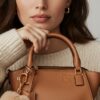 A close-up portrait of a woman with a natural and soft expression, wearing a cozy cream-colored knit sweater. She is holding a luxurious tan leather handbag inspired by the Loewe Hammock Bag, featuring gold zipper details and a structured design.