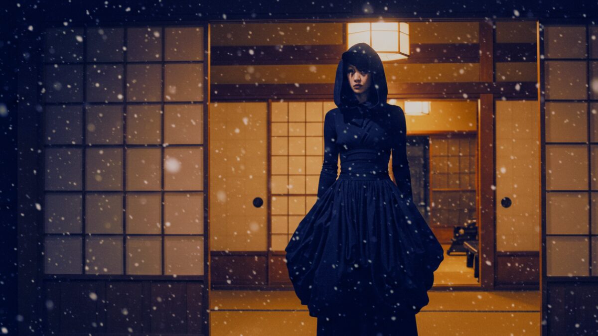 穏やかな雪が降る中、日本の伝統的な木造家屋の玄関に佇む、全身黒のアヴァンギャルドな衣装に身を包んだスタイリッシュな女性。ドラマチックなボリュームのある流れるような黒のロングドレスに、構築的な黒のトップスとユニークなフード付きのヘッドピースを重ねている。