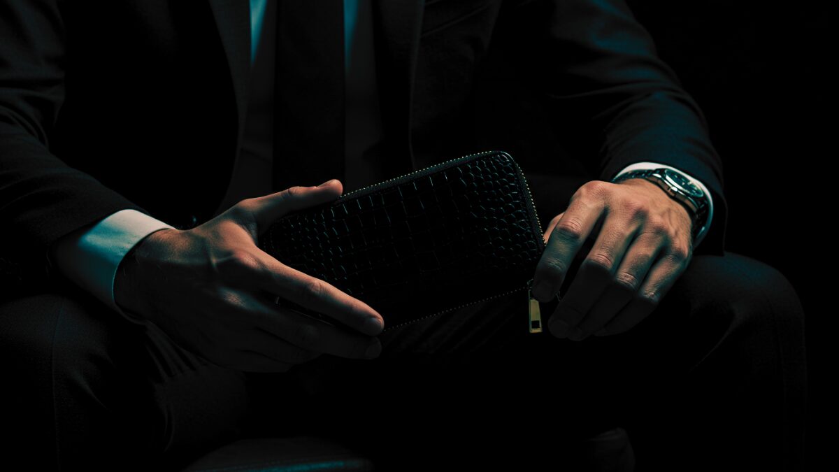 ダークテーラードスーツに白いドレスシャツを着た洗練された男性が、薄暗いエレガントな空間に座っている。手に持っているのは高級感のある黒のクロコダイル型押しレザーのファスナー付き財布で、ファスナーの引き手はゴールド。照明はムーディーかつドラマチックで、財布の光沢のある表面に微妙な反射を落としている。