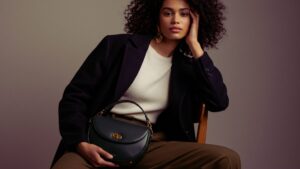 ダークカラーの巻き髪と落ち着いた表情が印象的なスタイリッシュな若い女性が、ミニマルでニュートラルトーンの背景の前で木製の椅子に座っている。肩にはオーバーサイズのウールコートを羽織り、白いノースリーブトップスとハイウエストのオリーブグリーンのパンツを着用。膝の上にはダークグリーンの三日月型レザーバッグ（ゴールドの金具付き）があり、片手でカジュアルに持っている。