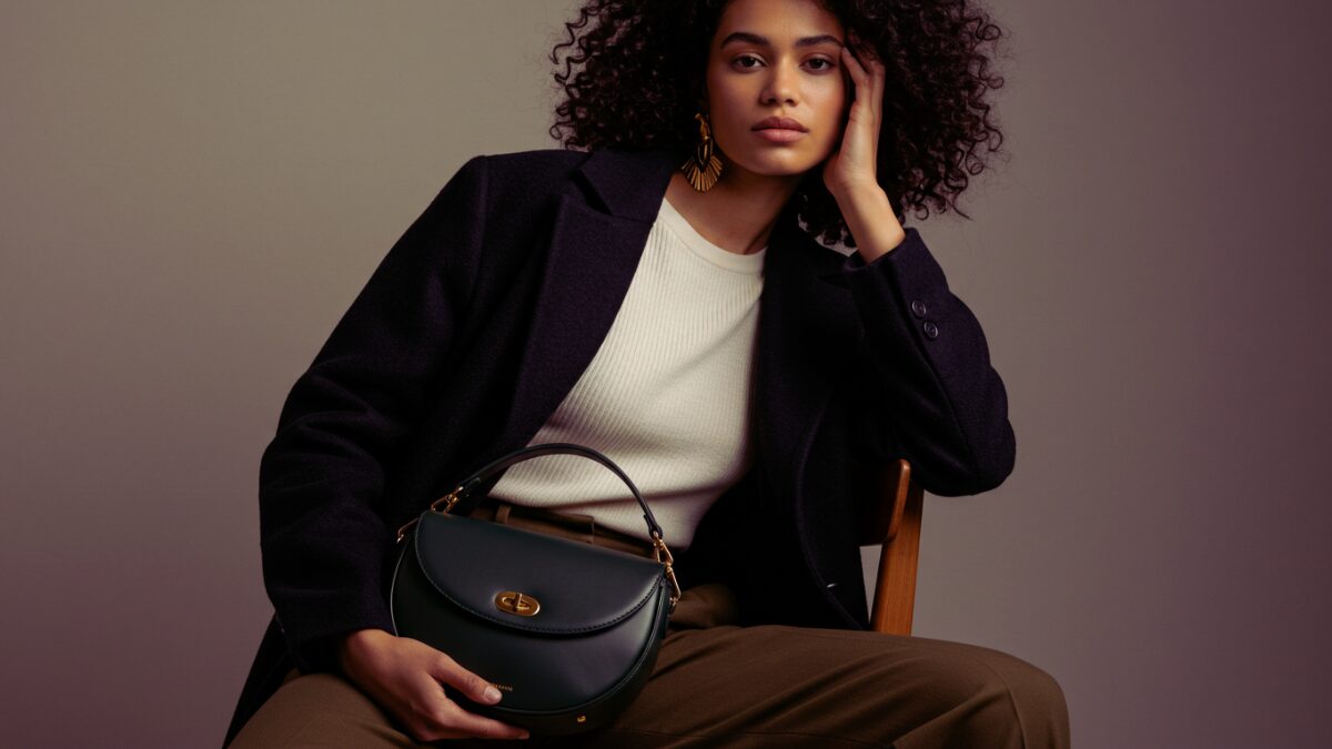 ダークカラーの巻き髪と落ち着いた表情が印象的なスタイリッシュな若い女性が、ミニマルでニュートラルトーンの背景の前で木製の椅子に座っている。肩にはオーバーサイズのウールコートを羽織り、白いノースリーブトップスとハイウエストのオリーブグリーンのパンツを着用。膝の上にはダークグリーンの三日月型レザーバッグ（ゴールドの金具付き）があり、片手でカジュアルに持っている。