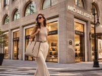 マイケル・コース風の高級感ある店舗前を、ベージュ系のきれいめコーデを着た女性がバッグを持って歩く都会的なファッションビジュアル。